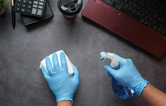 Person wearing blue gloves cleaning a worktop desk with a spray bottle and cloth near a laptop and calculator